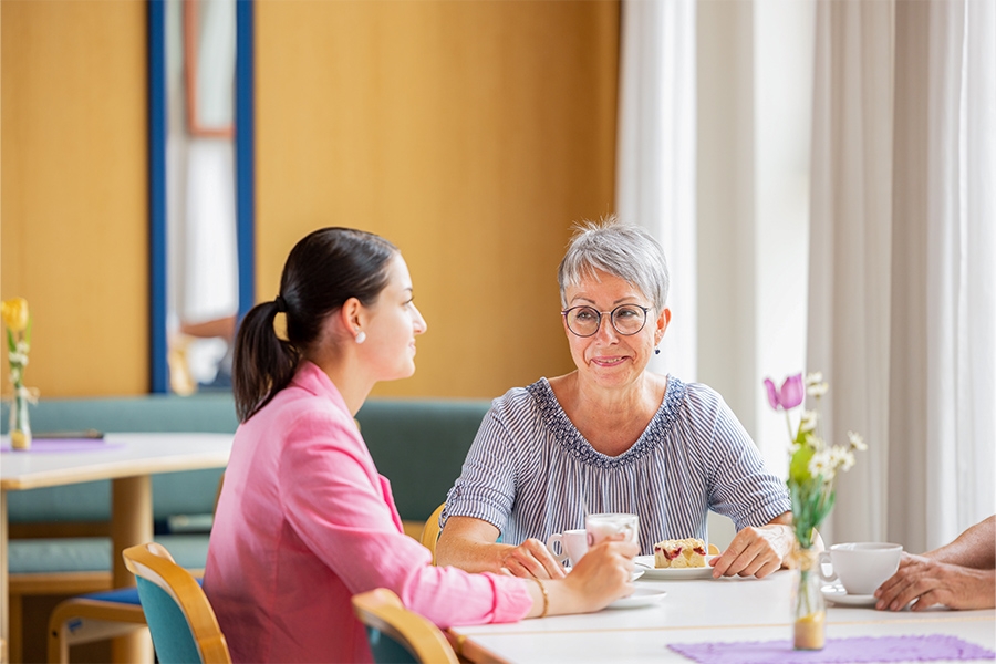 Zwei Frauen sitzen an einem Tisch, unterhalten sich, trinken Kaffee und essen Kuchen.