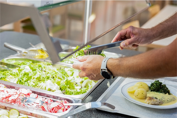 Jemand befüllt seine Schüssel am Salatbuffet. Vor ihm steht ein Teller mit einem Fleischgericht mit Kartoffeln. Jemand befüllt seine Schüssel am Salatbuffet. Vor ihm steht ein Teller mit einem Fleischgericht mit Kartoffeln.
