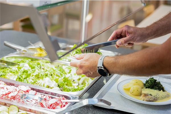 Jemand befüllt seine Schüssel am Salatbuffet. Vor ihm steht ein Teller mit einem Fleischgericht mit Kartoffeln.