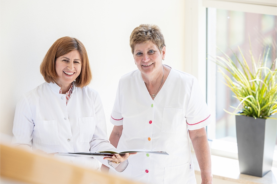 Zwei Mitarbeiterinnen der Klinik stehen nebeneinander und lächeln in die Kamera. Eine der Frauen hält eine Akte.