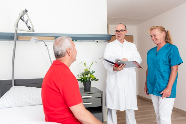 Ein Patient sitzt in seinem Zimmer auf seinem Bett und unterhält sich mit einem Arzt und einer Frau vom Pflegepersonal, die ihm gegenüber stehen. Ein Patient sitzt in seinem Zimmer auf seinem Bett und unterhält sich mit einem Arzt und einer Frau vom Pflegepersonal, die ihm gegenüber stehen.