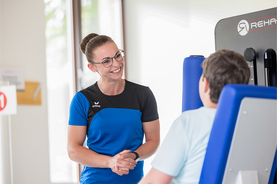 Eine Therapeutin lächelt eine Patientin an, die auf einem Trainingsgerät sitzt.