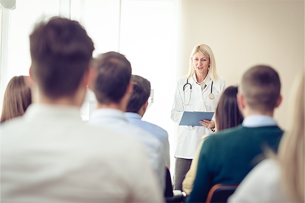 Mehrere Menschen hören dem Vortrag einer Ärztin zu. Mehrere Menschen hören dem Vortrag einer Ärztin zu.